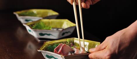 A person holding chopsticks over a small dish with food, likely sushi, on a wooden table.