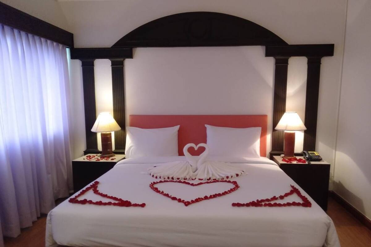 A hotel room with a large bed, two bedside lamps, and a heart-shaped decoration made of rose petals on the bed.