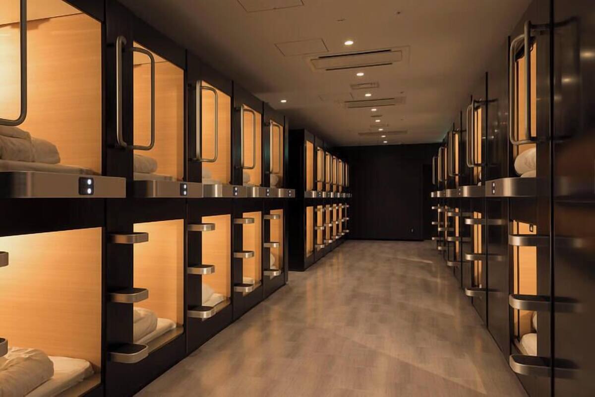 Une rangée de lits superposés, chacun équipé d’un éclairage individuel, un plancher en bois et un mur sombre.