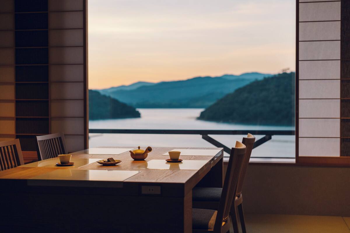 Een eettafel met stoelen, een raam met uitzicht op een meer en bergen, en een theepot en kopjes op de tafel.