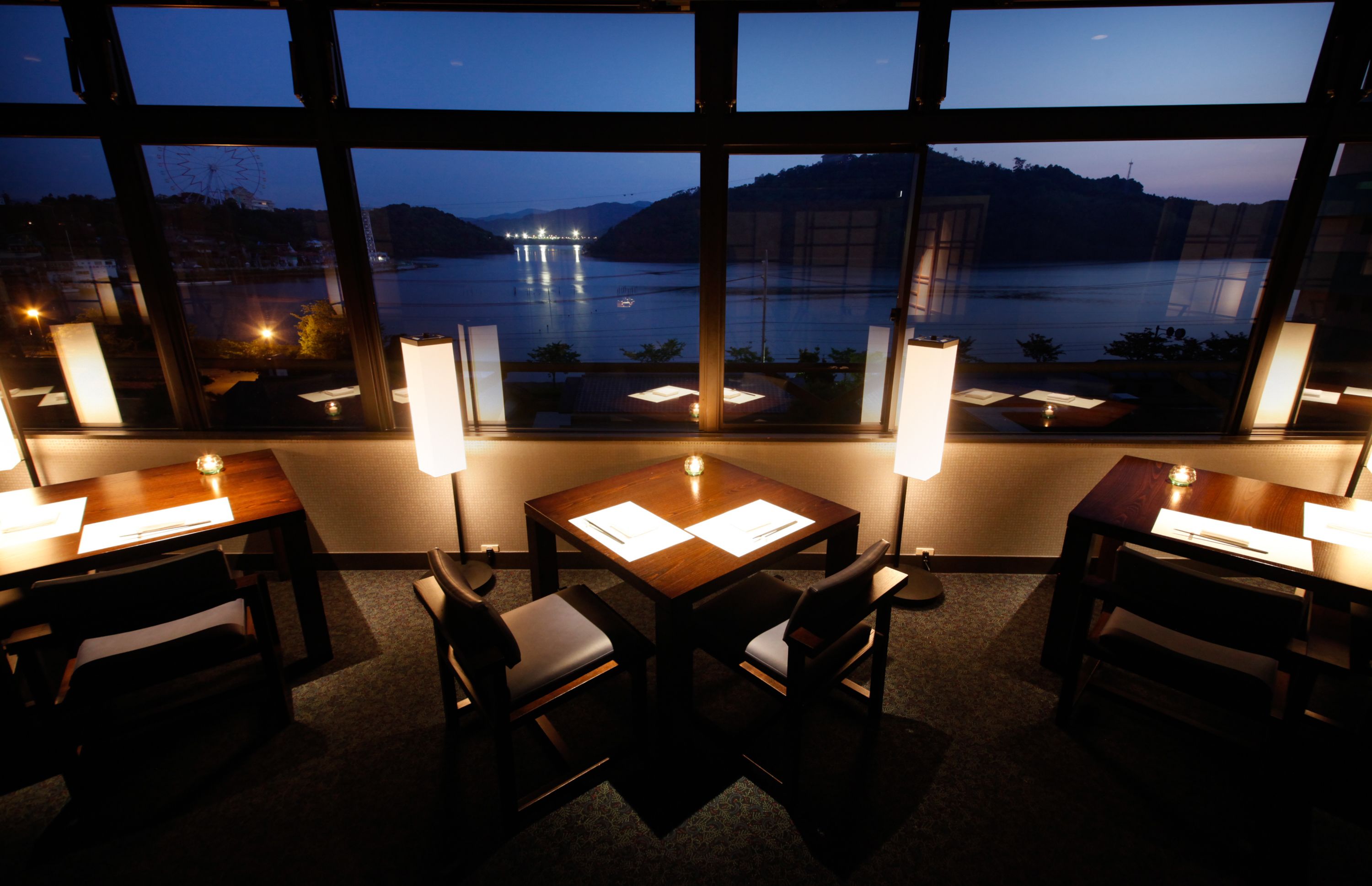 A dining area with a view of a lake at dusk, featuring tables with menus and candlelit lamps.