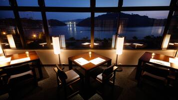 A dining area with a view of a lake at dusk, featuring tables with menus and candlelit lamps.