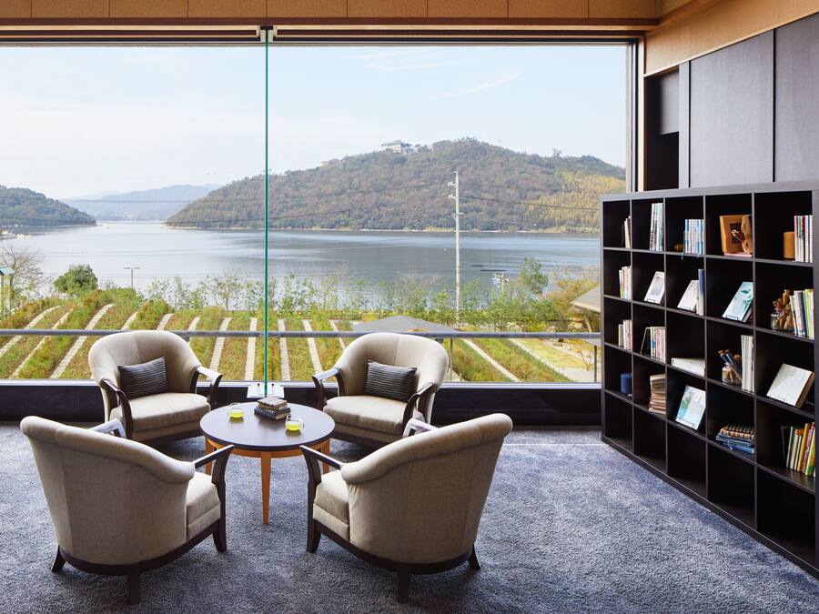A room with a large window overlooking a lake and hills, two armchairs, a small round table, and a bookshelf.