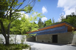 A modern building with a concrete facade and a wooden roof, surrounded by trees and a clear sky.
