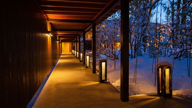 Un corridoio con pareti e colonne in legno, illuminato da luci calde, che conduce a un'area esterna innevata con alberi.