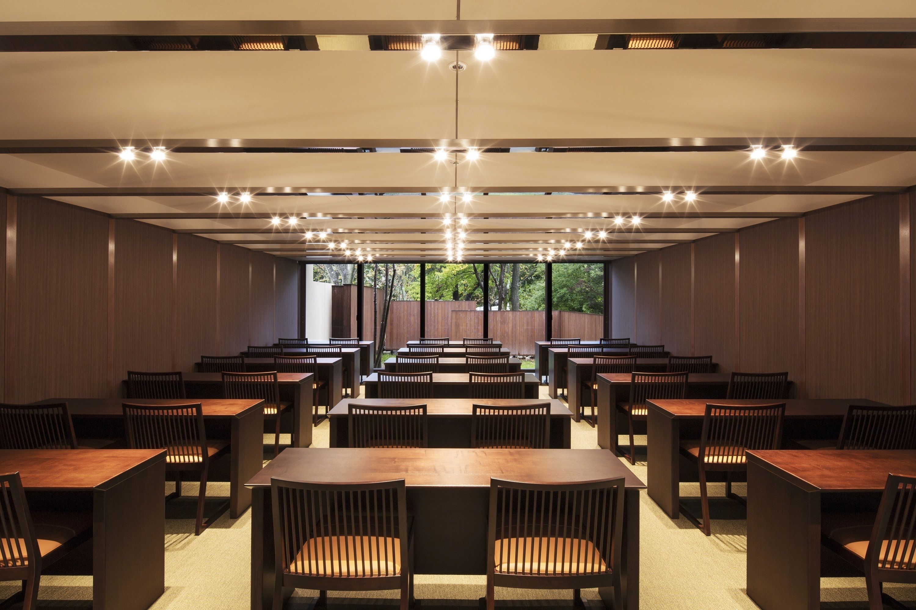 A spacious dining area with multiple tables and chairs, a high ceiling with recessed lighting, and large windows providing a view of the outdoors.