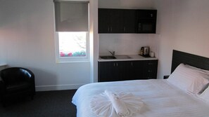 A hotel room with a bed, a small kitchenette, a window with a view of flowers, and a chair.