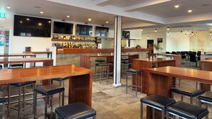 A restaurant with wooden tables and black bar stools, a bar area with shelves of bottles, and a large TV screen. - Queens Head Hotel (North Hobart)