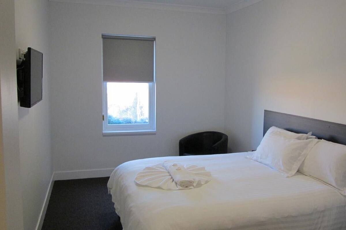 A hotel room with a single bed, a window with blinds, a chair, and a flat-screen TV mounted on the wall.