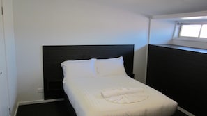 A single bed with white bedding and pillows, a dark headboard, and a small window above the bed.