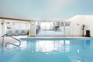 An indoor swimming pool with clear glass walls, a pool ladder, and a view of a snowy outdoor area.