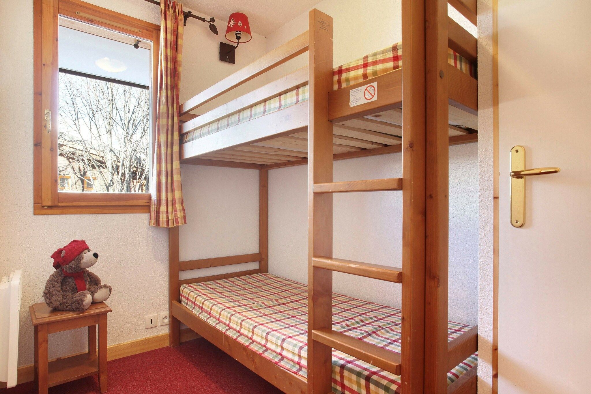 Ein Schlafzimmer mit Etagenbett und Fenster, ein Teddybär auf einem kleinen Tisch und eine Tür mit Griff.