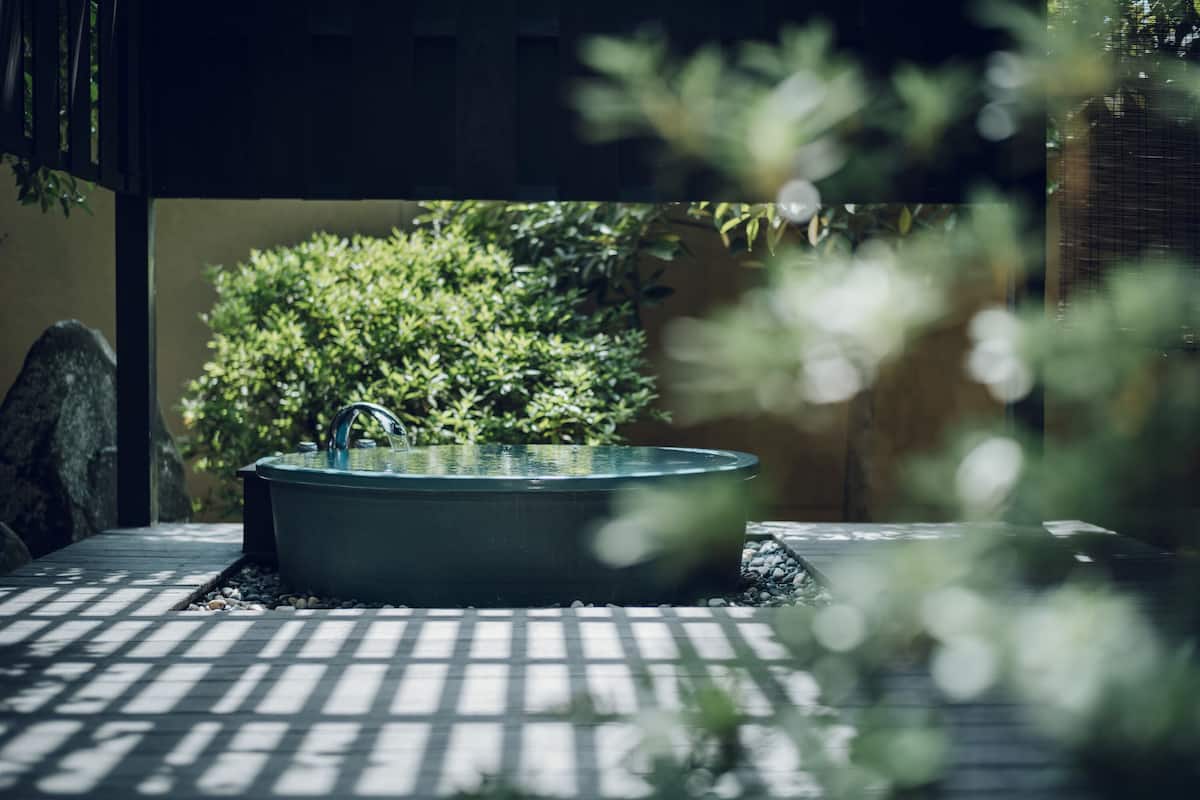 Une baignoire extérieure rectangulaire, dotée d’un robinet, entourée de galets et de plantes, et située sous une pergola dont le sol est recouvert de carreaux.