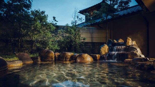 Un jardín japonés tradicional con un arroyo cristalino, faroles de piedra y un edificio de madera con tejado de tejas.