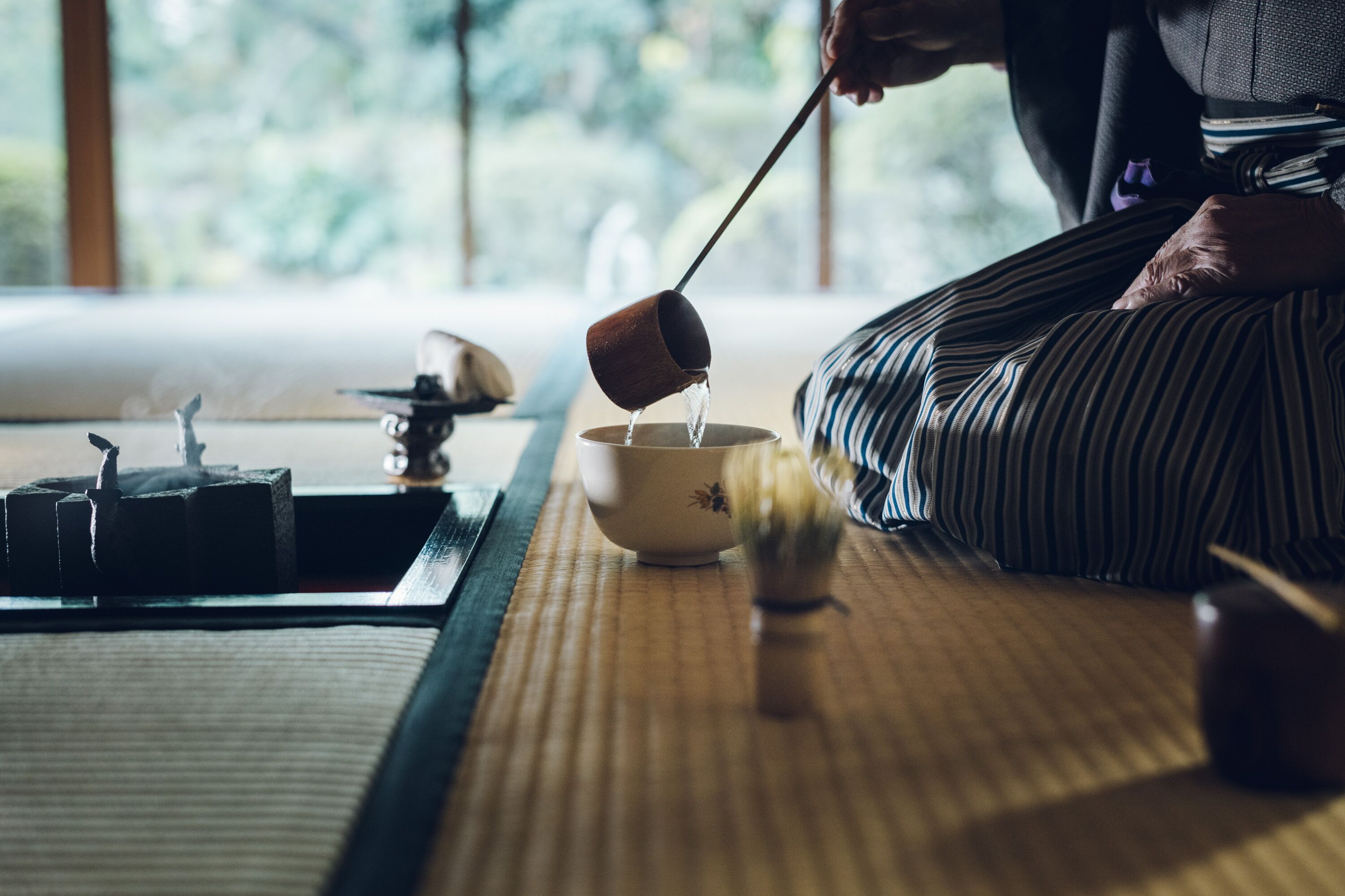 Een persoon zit op een tatami mat en giet water uit een kop in een kom.