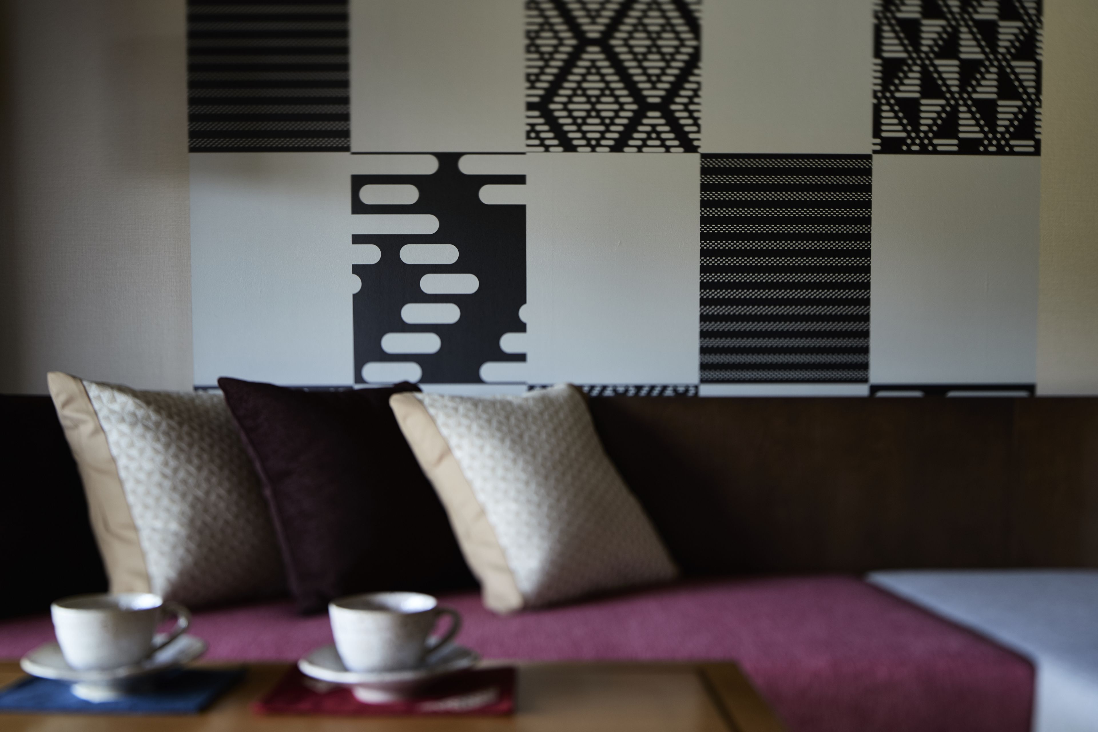 Un área de estar con sofá y cojines, una mesa con una taza y plato, y una pared con patrones geométricos.