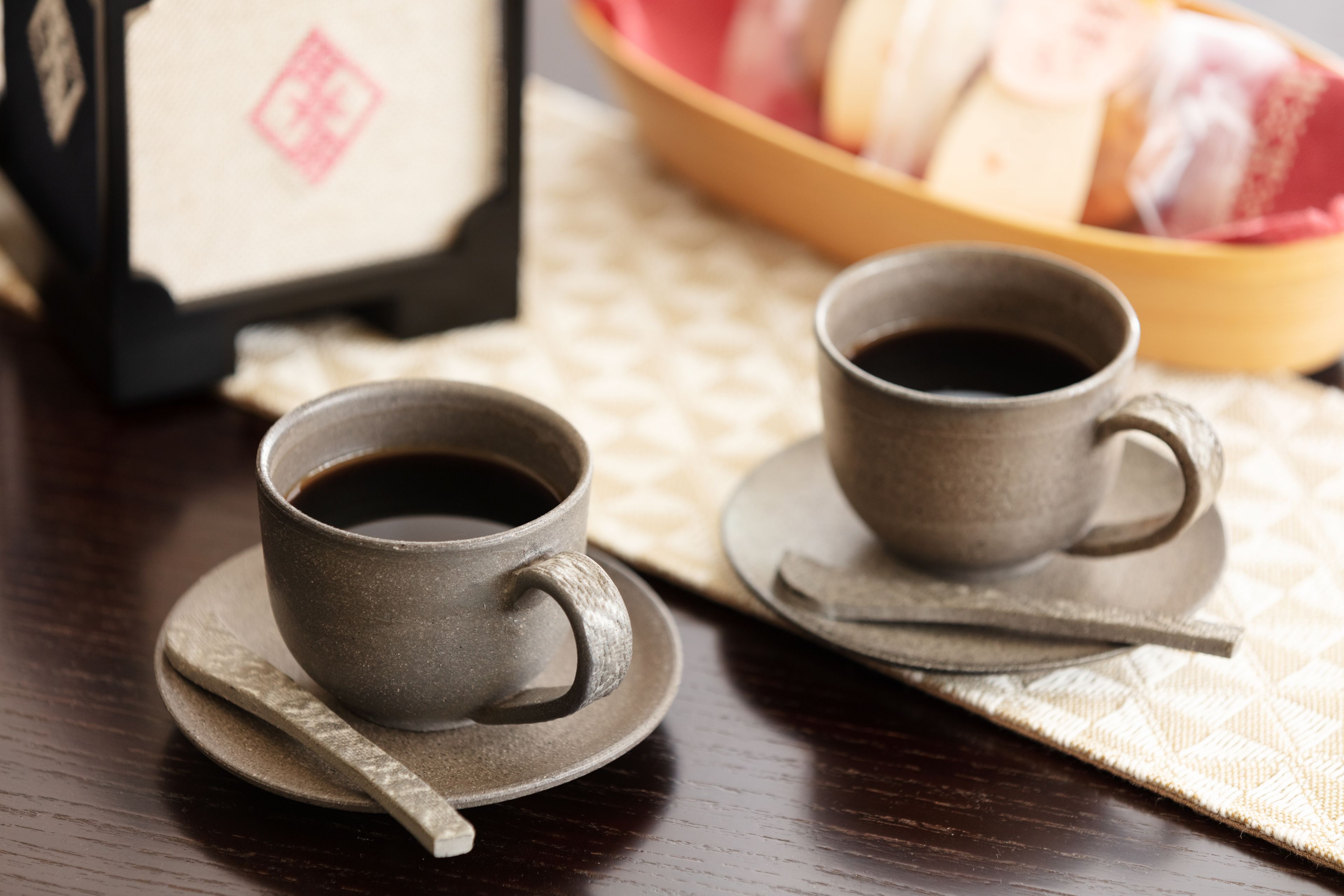 Two ceramic cups of coffee on saucers with spoons, a wooden tray, and a basket of pastries in the background.