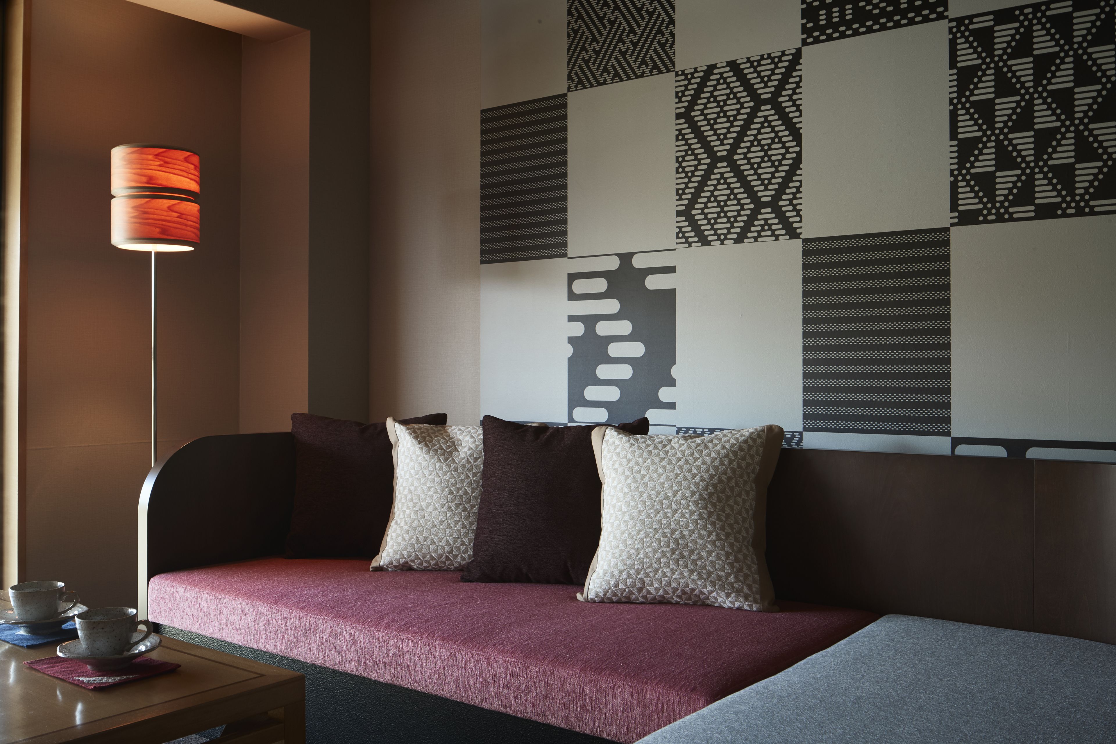 A modern hotel room with a sofa, pillows, a table with cups, and a patterned wall.