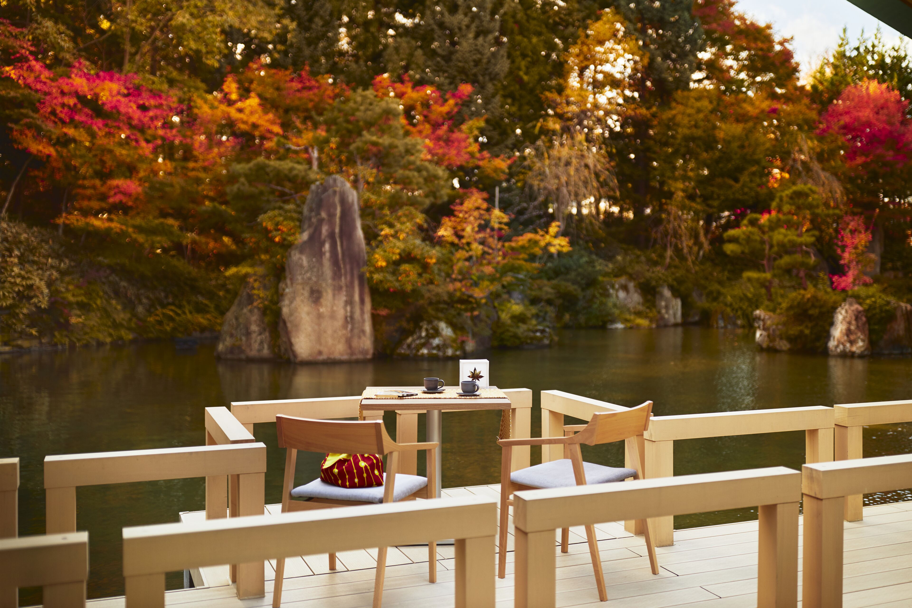 Una terraza de madera con sillas y una mesa, con vista a un estanque rodeado de árboles de colores otoñales.