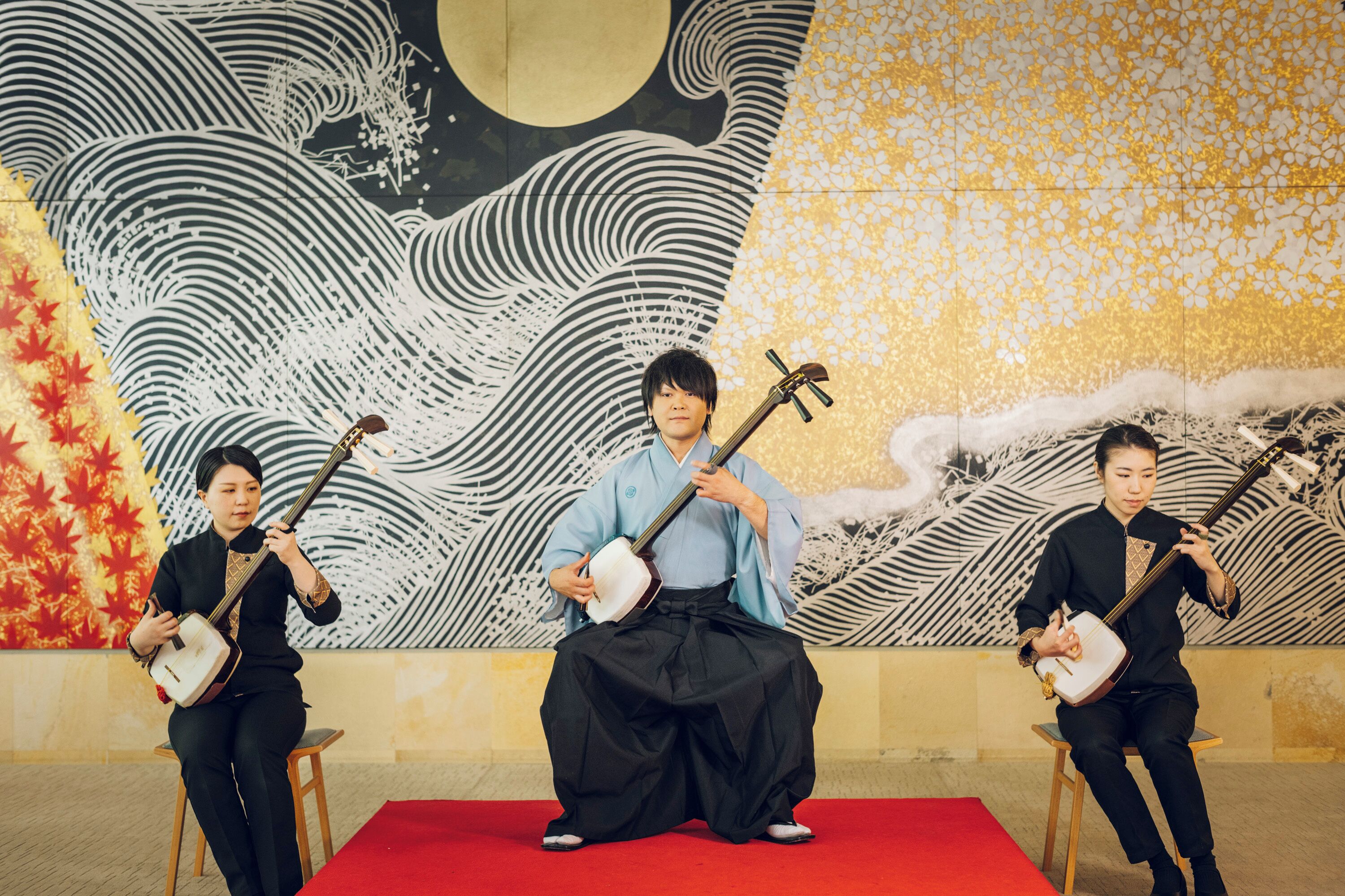Three musicians seated on stools on a red carpet, playing traditional string instruments.