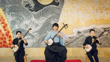 Three musicians seated on stools on a red carpet, playing traditional string instruments.