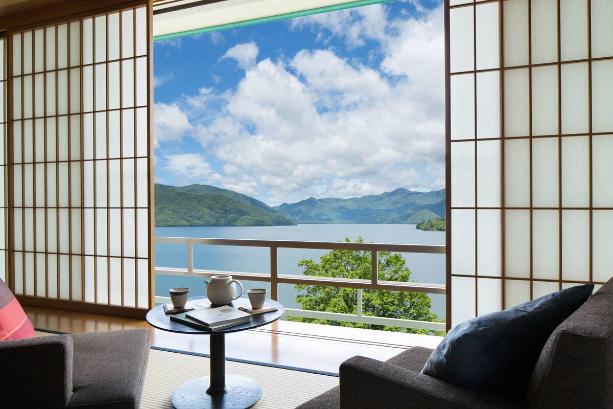 A room with tatami flooring, a low table with a teapot and cups, and a sofa with cushions. A large window offers a view of a lake and mountains.