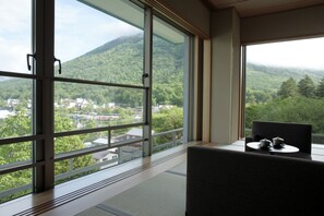 Una habitación con un ventanal que ofrece vistas a una montaña boscosa y un pueblo. Hay una silla y una mesita con tetera y taza.