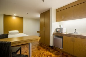 Habitación de hotel compacta con una cama, un pequeño rincón de cocina, zona de comedor con mesa y sillas, y suelo de madera.