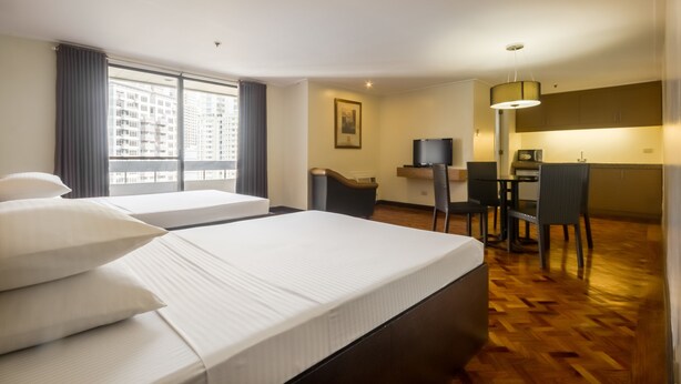 A hotel room with two beds, a desk, a chair, a television, and a view of buildings through the window. - Salcedo Suites (Makati)