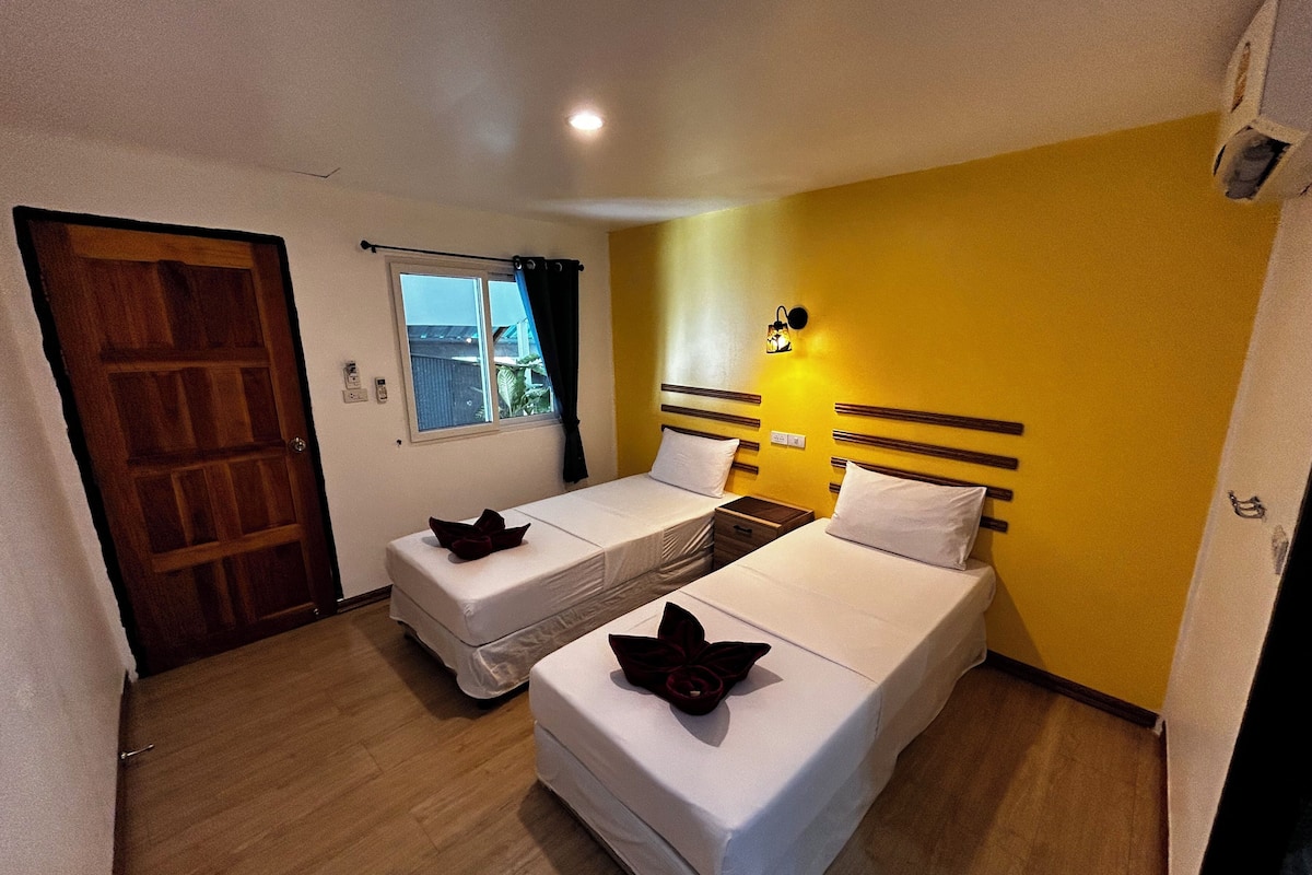 Una habitación con dos camas individuales, una puerta de madera, una ventana con cortinas y una lámpara fijada a la pared.