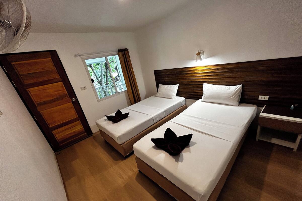 Habitación de hotel con dos camas, cabecero de madera, ventana con cortinas, mesita de noche y un ventilador.