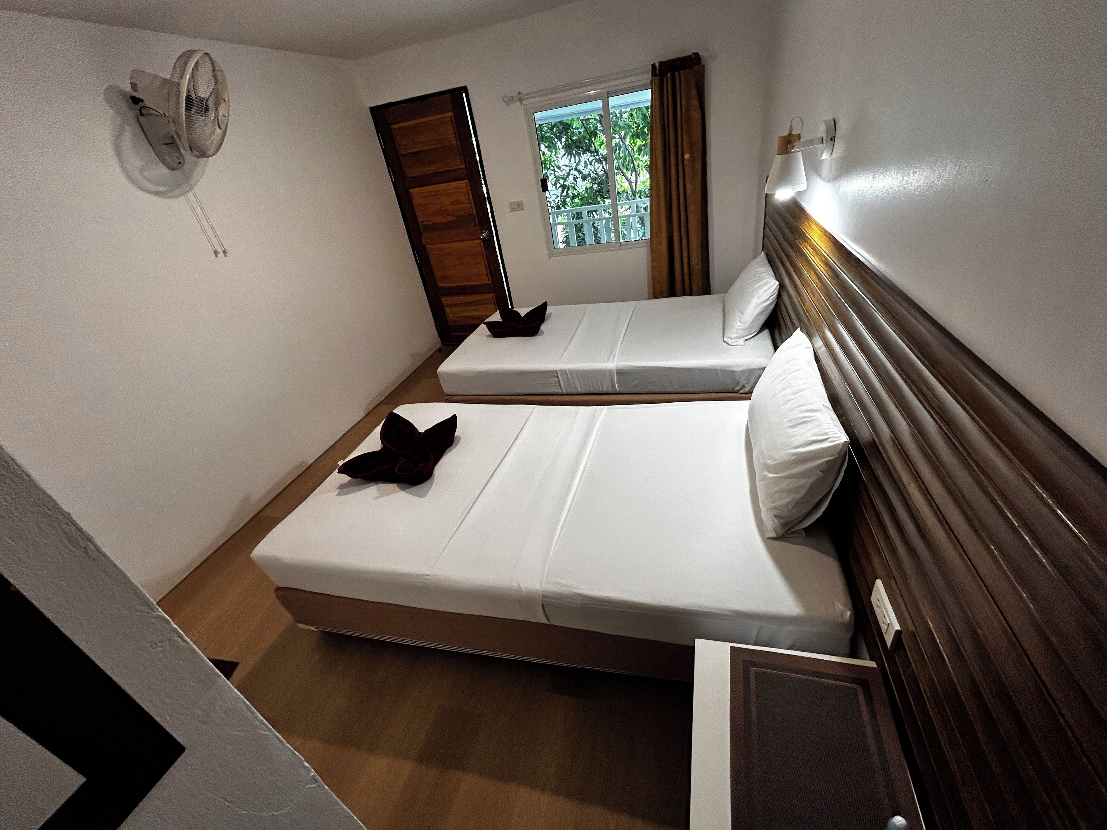 A small, compact hotel room with two single beds, a window with curtains, a wall-mounted fan, and a wooden headboard.