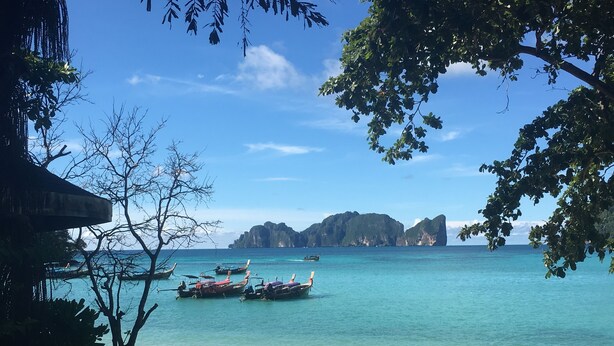 A beach with clear turquoise water, boats, and rocky islands in the background. - Coco's Guesthouse (Ko Phi Phi)