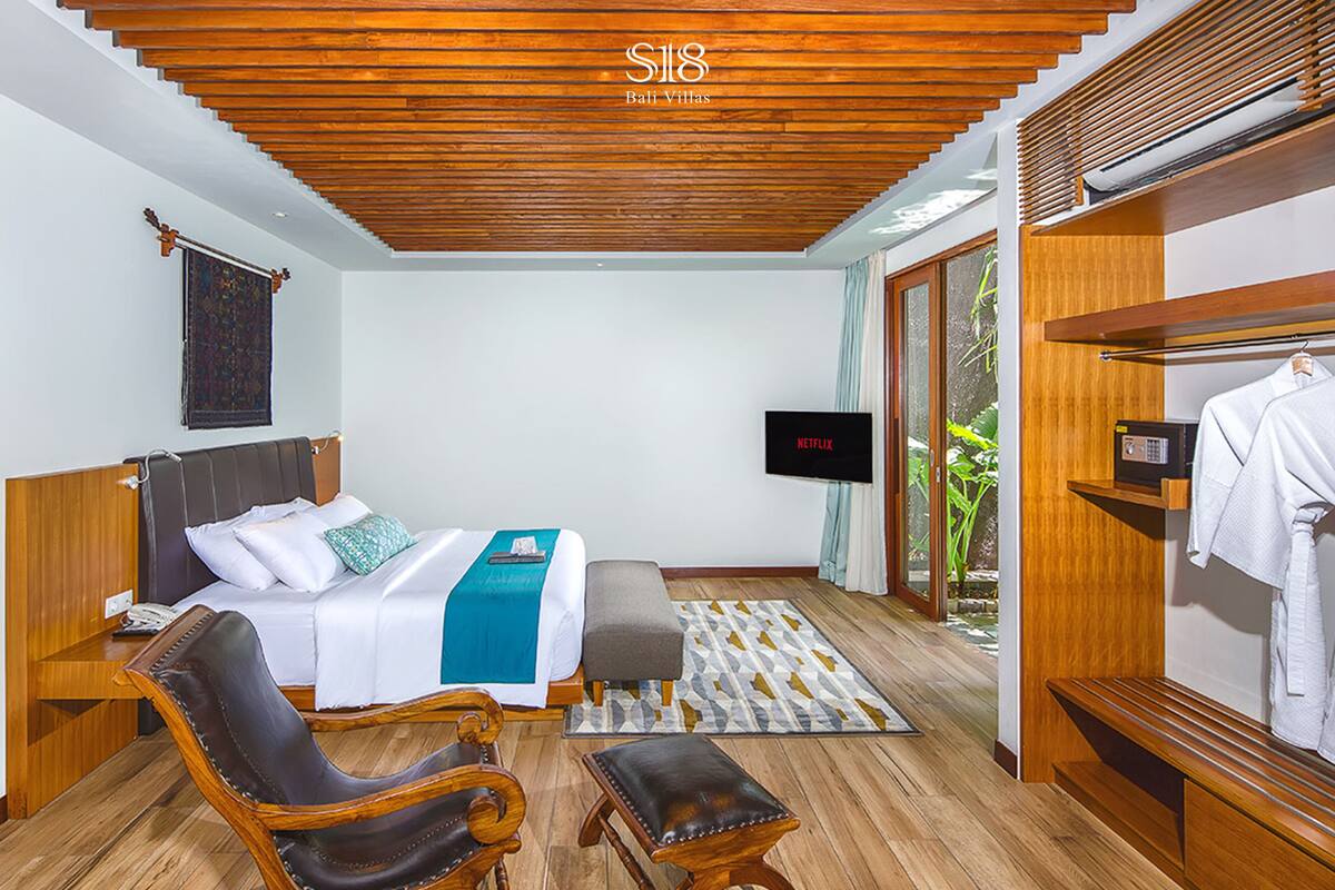 Une chambre d’hôtel moderne avec un plafond en bois, un lit recouvert d’une couverture bleue, une télévision, une armoire en bois et un coin salon comprenant un fauteuil et un pouf.
