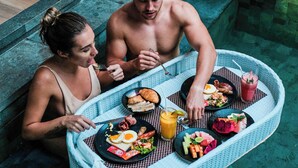 Una pareja cena junto a una piscina, con una bandeja de comida y bebidas.