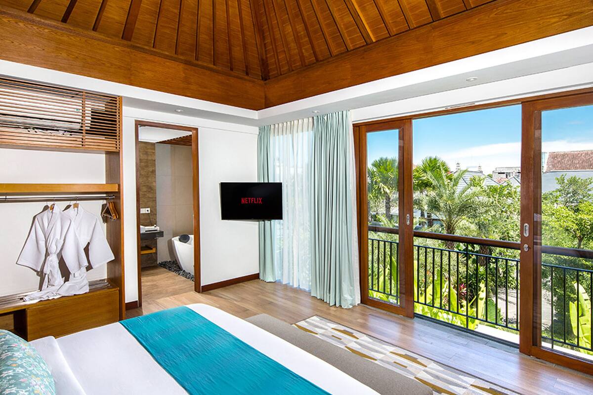 Une chambre d’hôtel moderne avec un grand lit, un téléviseur fixé au mur, un balcon donnant sur un paysage verdoyant et un plafond en bois.
