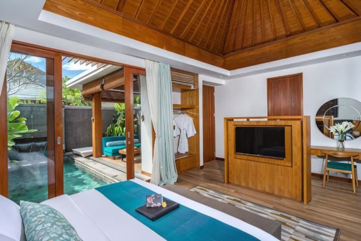 A bedroom with a large bed, a TV, a desk, and a view of an outdoor pool.