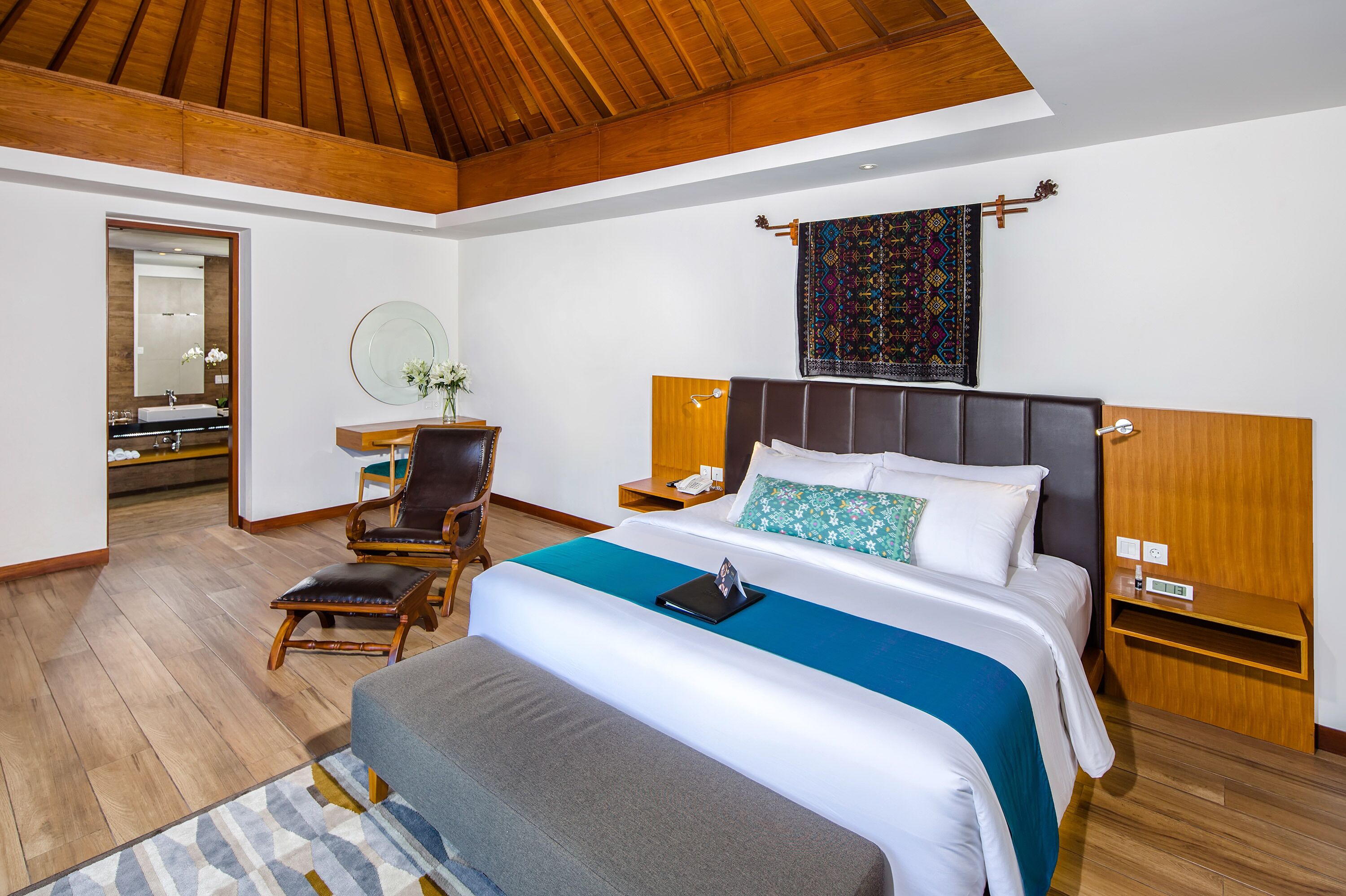 A modern hotel room with a wooden ceiling, a large bed with a blue accent, a wooden nightstand, a chair, and a small desk with a mirror.