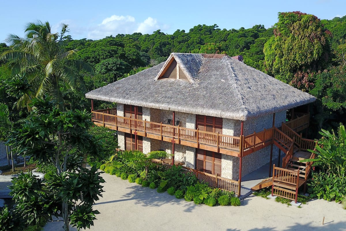 A two-story building with a thatched roof, surrounded by lush greenery and a sandy beach.