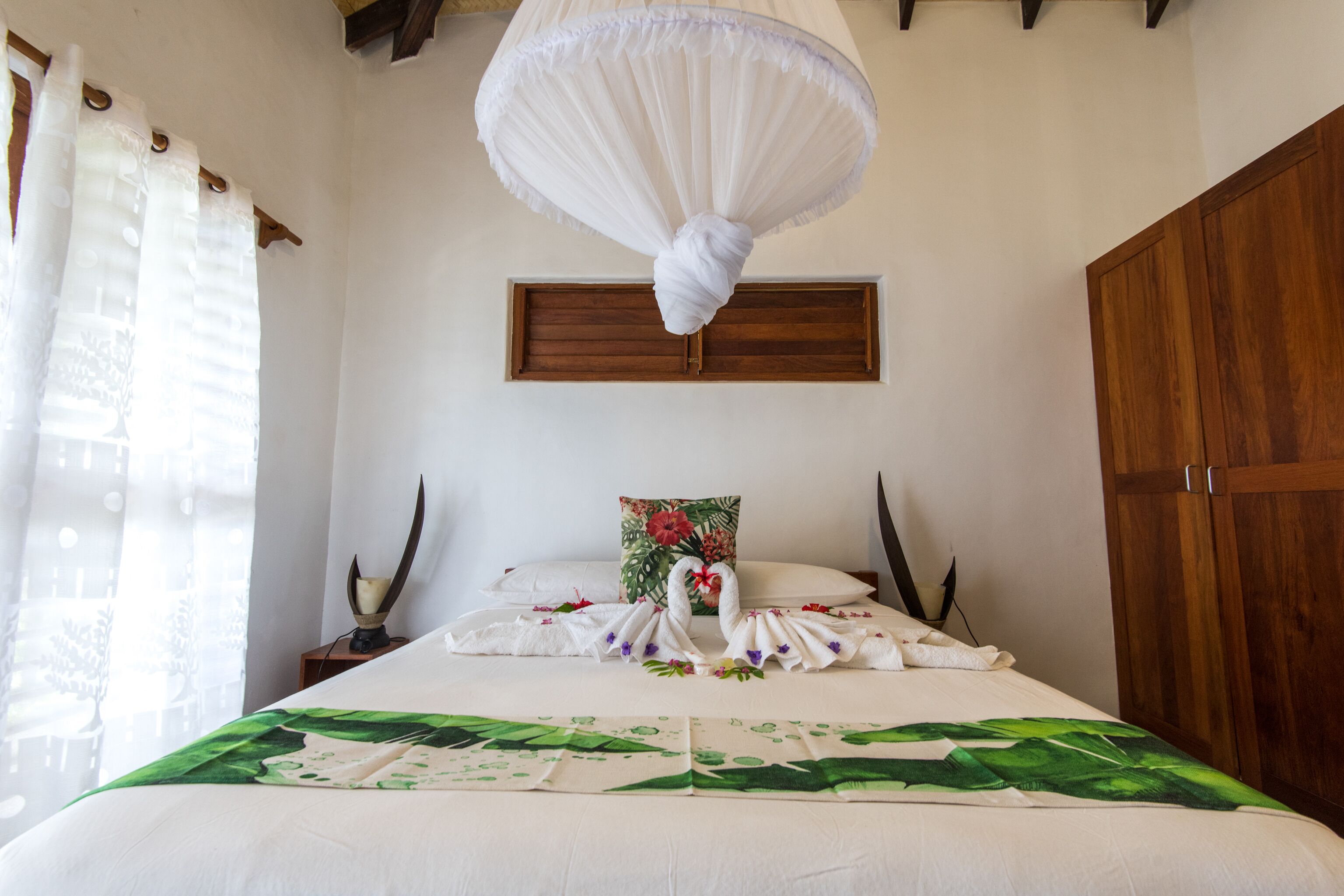 A neatly made bed with a towel swan centerpiece, a wooden headboard, a nightstand with a candle, and a large window with sheer curtains.