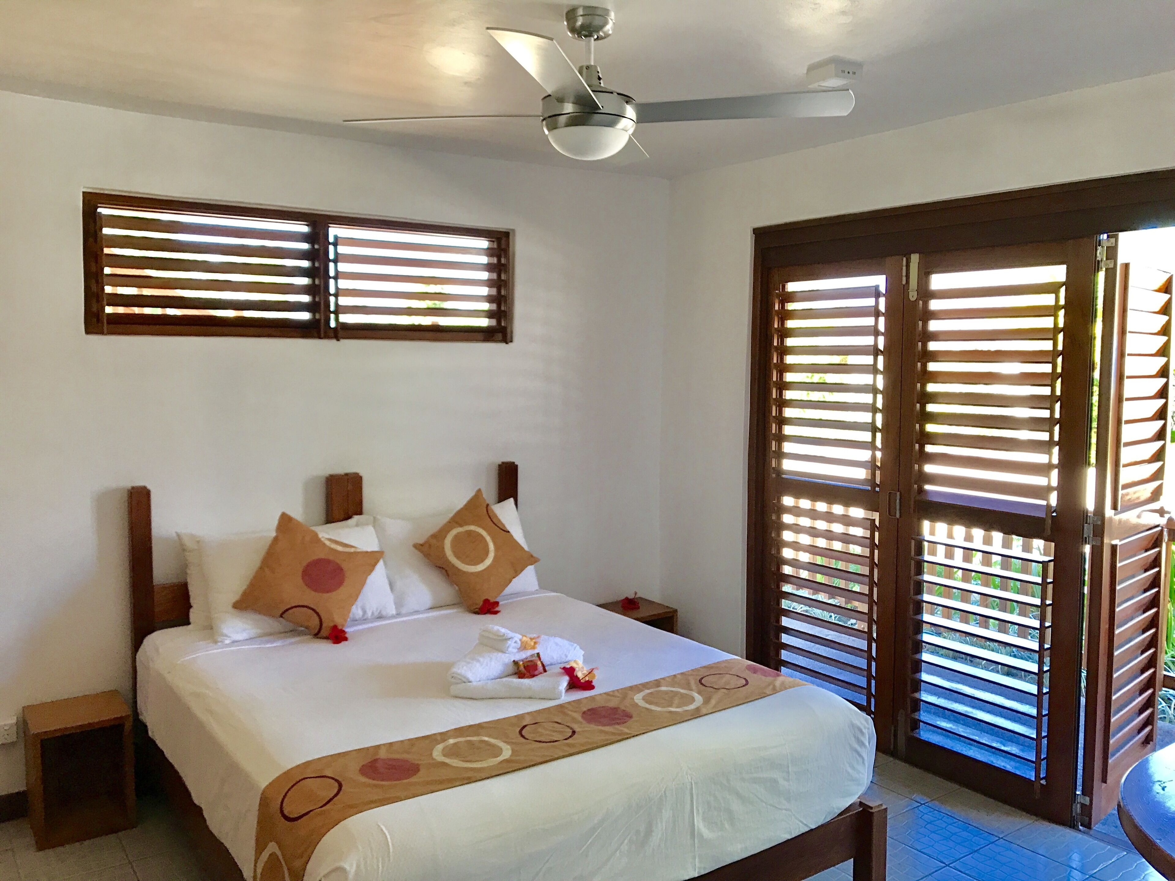 Quarto de hotel com cama, ventilador de teto e janela com persianas.