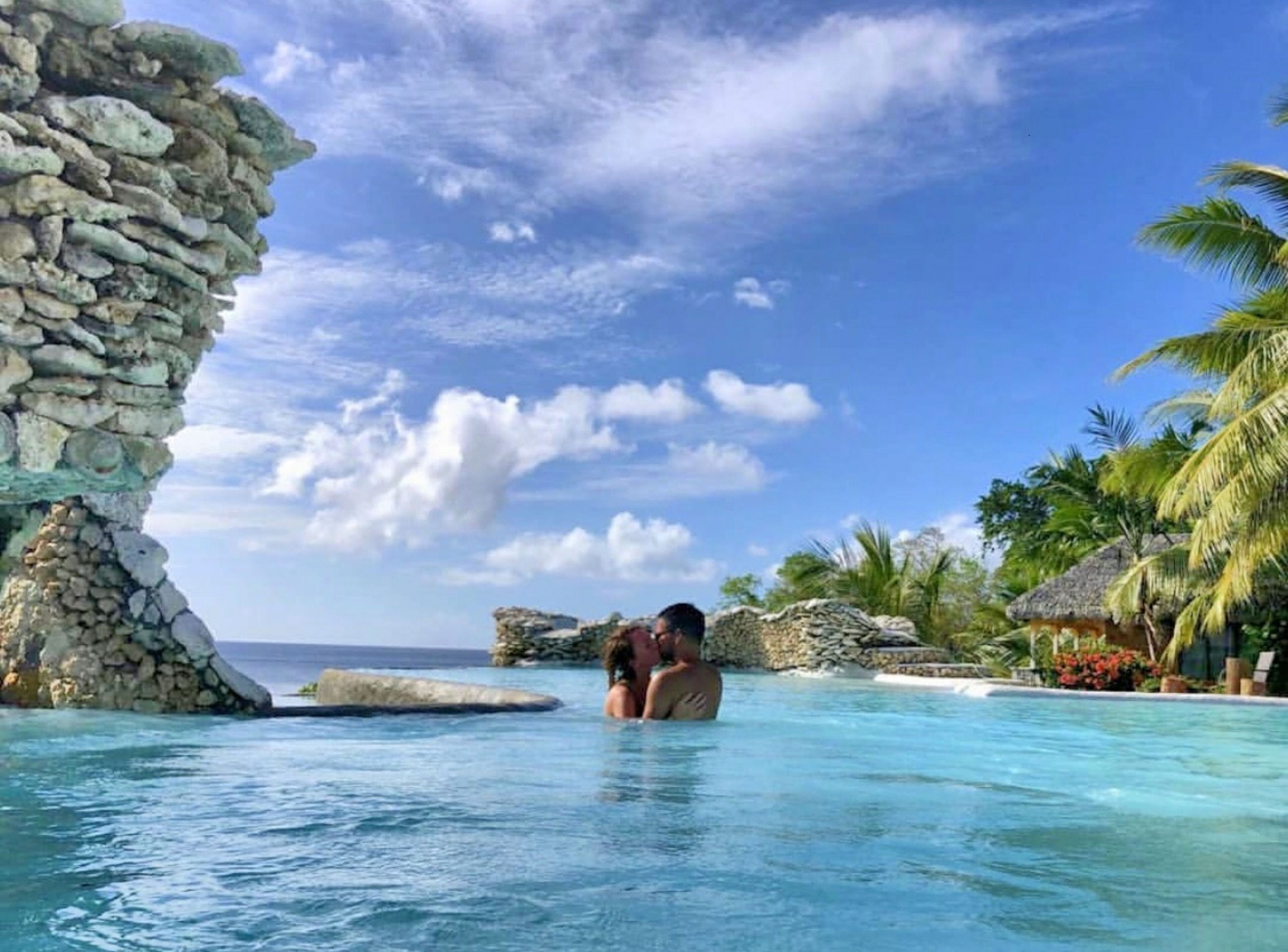 A couple in a clear blue pool with a stone rock formation and palm trees in the background.