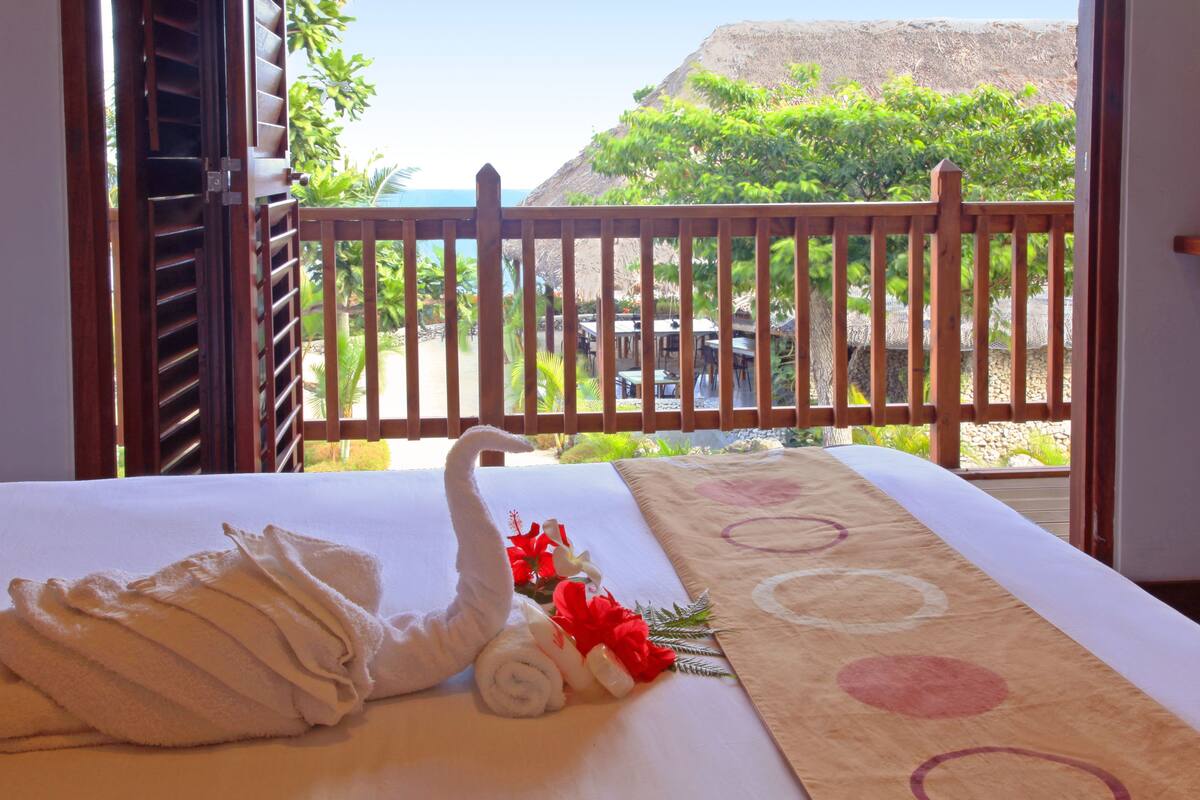 A hotel room with a bed, a wooden balcony railing, and a view of a mountain and greenery.