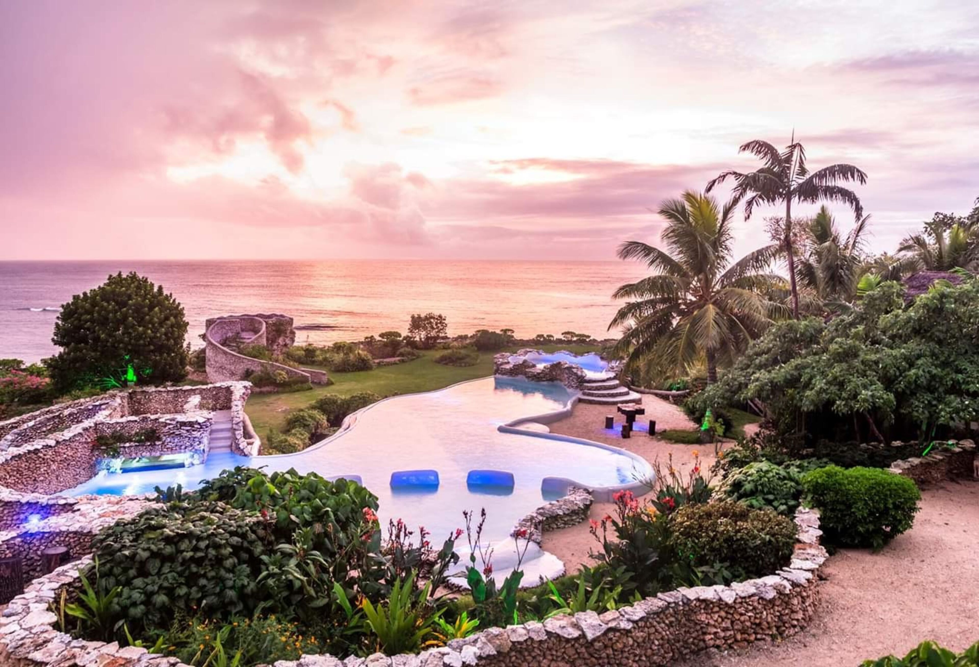 A pool area with a curved design, surrounded by tropical plants and palm trees, overlooking the ocean.