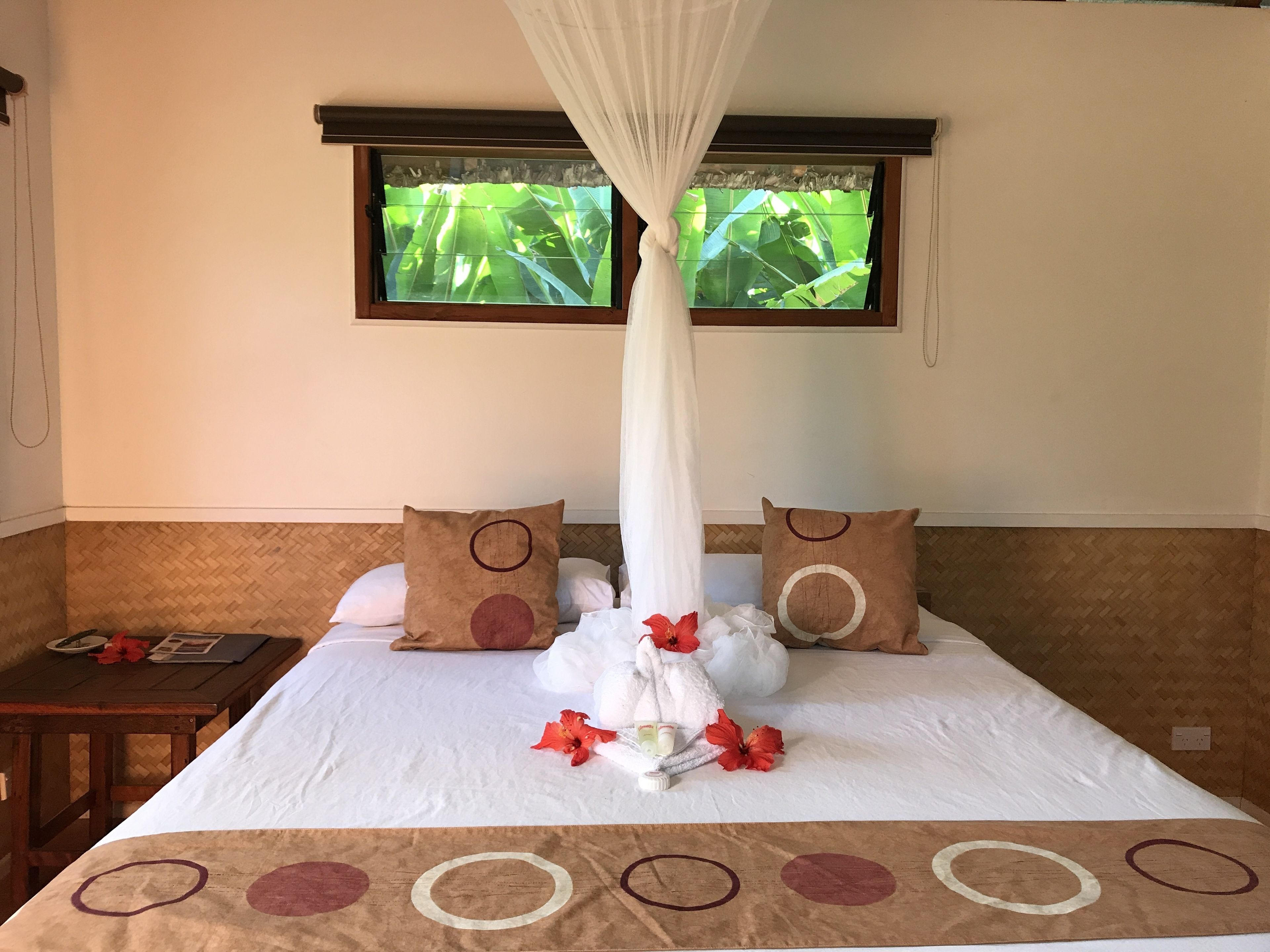 A hotel room with a bed, a window with a view of greenery, a bedside table with a lamp, and a curtain.