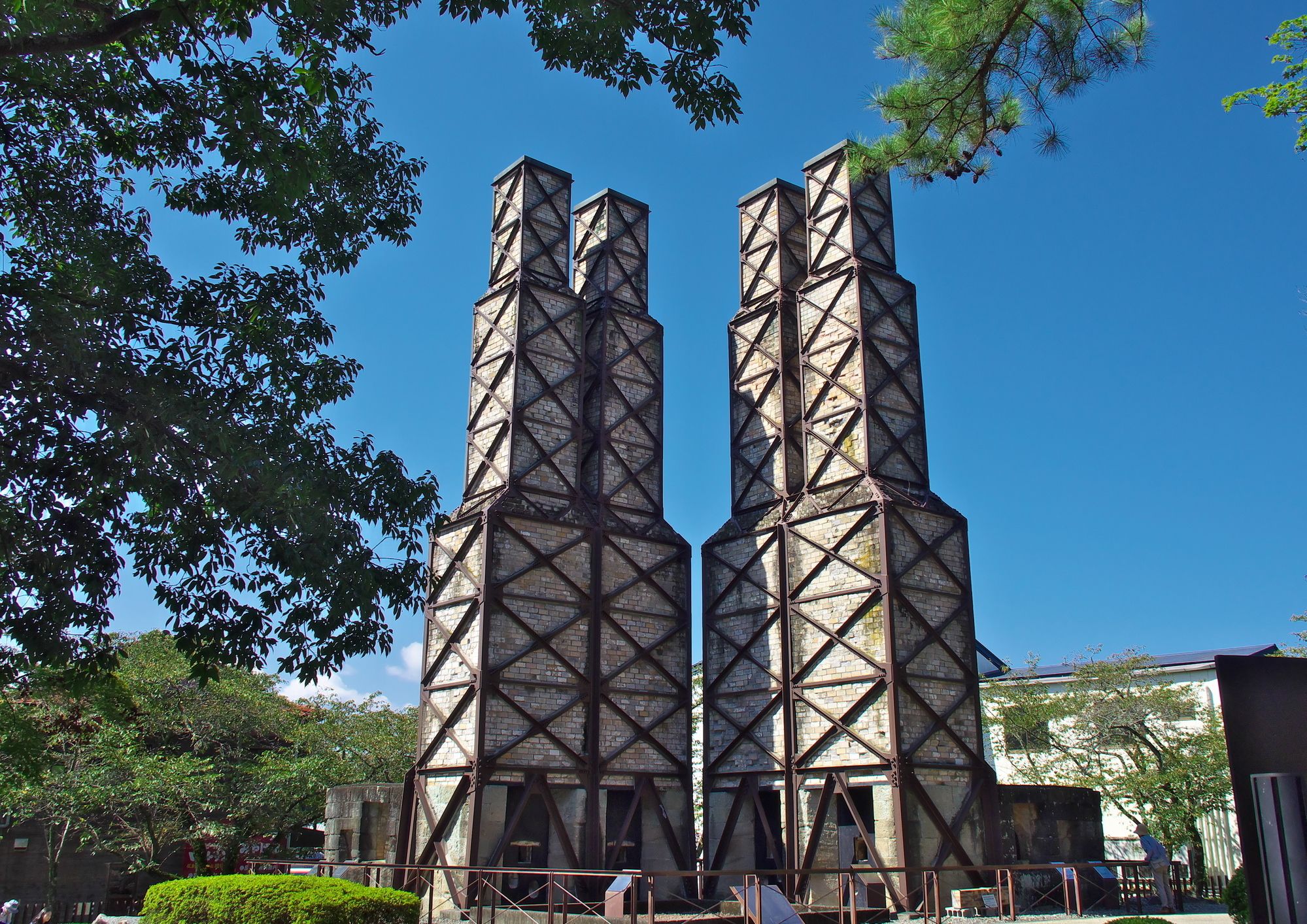 Two tall, industrial-style towers with a lattice framework, surrounded by trees and a clear blue sky.
