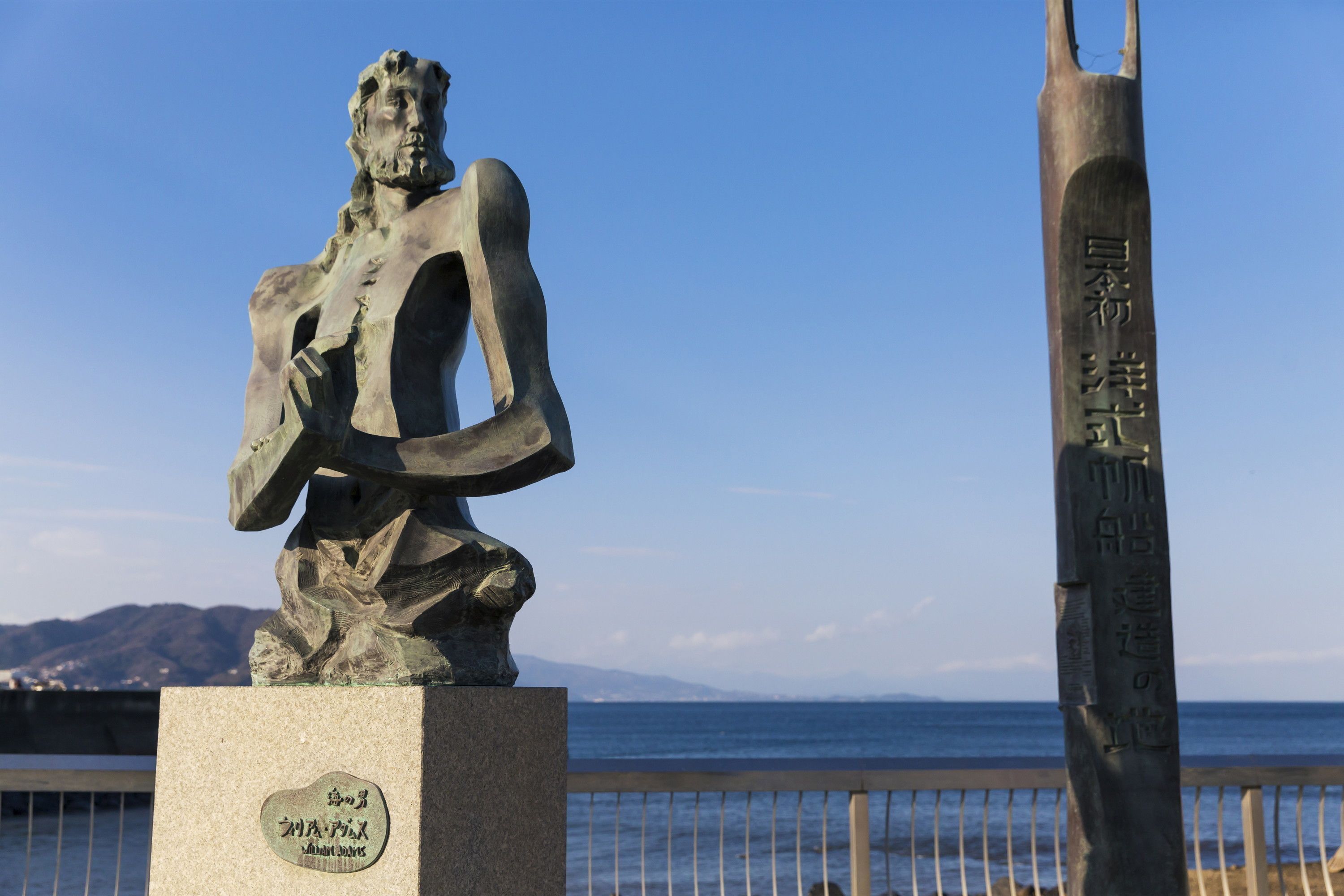 Une statue d’une personnalité historique, avec une plaque commémorative devant un paysage côtier.