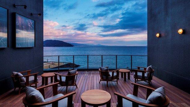 A wooden deck with round tables and chairs, overlooking a body of water with a view of distant land and a cloudy sky. - Hoshino Resorts KAI Anjin (Ito)