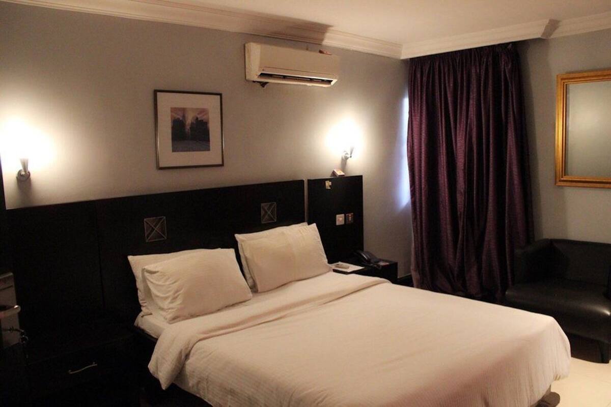 A hotel room with a large bed, a nightstand, a desk, a chair, an air conditioning unit, and a framed picture on the wall.
