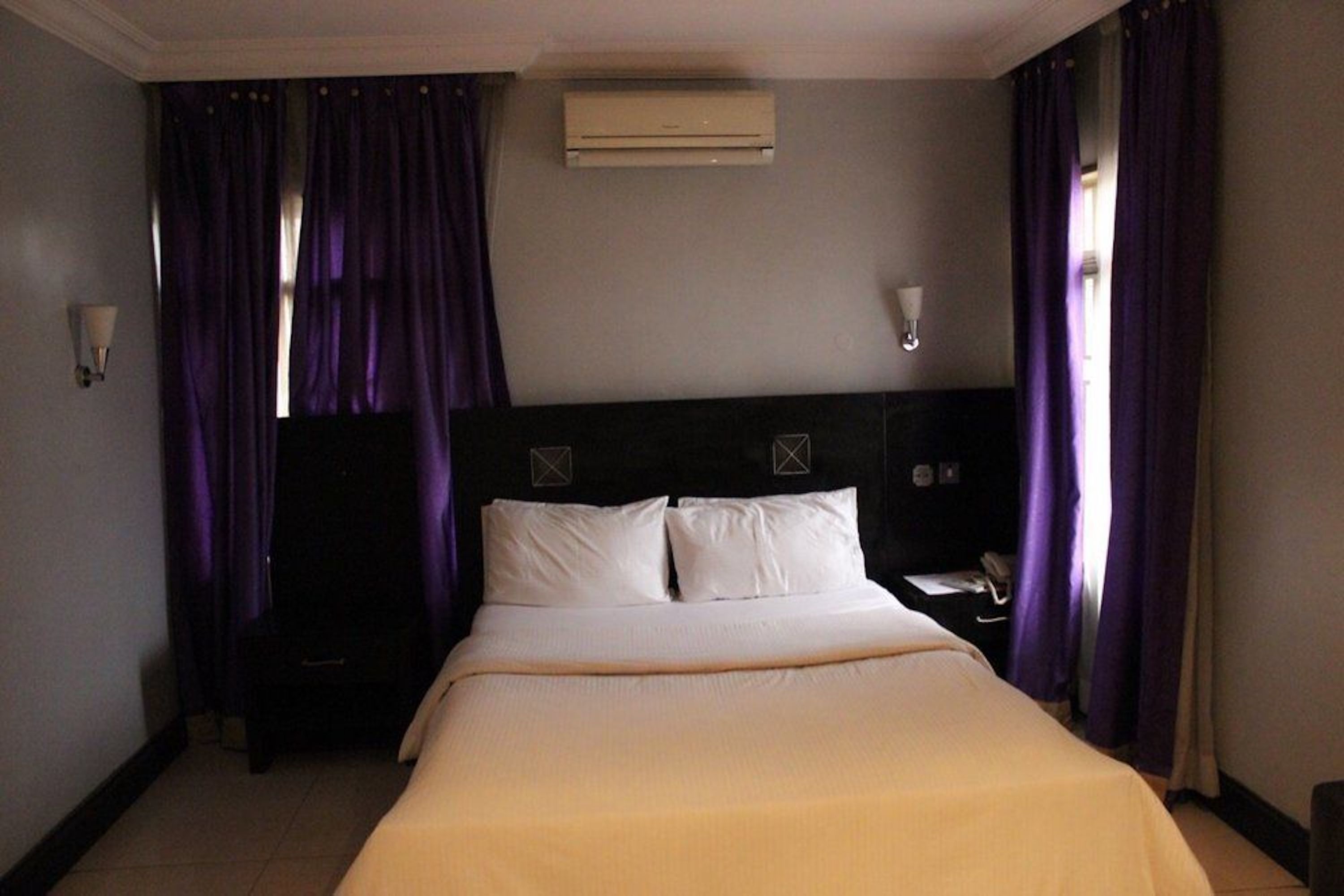 A hotel room with a large bed, a nightstand, an air conditioning unit, and purple curtains.